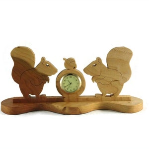 May include: Wooden squirrel clock with two squirrels flanking a round clock face. The squirrels are carved in a standing position with their tails curled up. The clock face has black Roman numeral markings and black hands.
