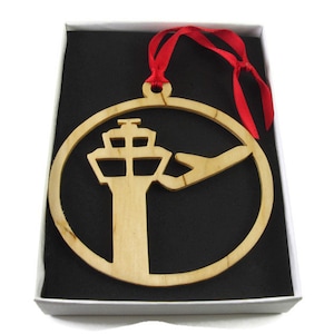 May include: A wooden Christmas ornament shaped like a circle with a cut-out design of an airport control tower and an airplane. The ornament is hanging from a red ribbon and is displayed in a white box with a black interior.