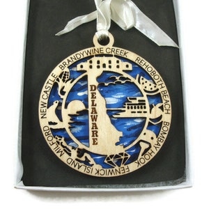 May include: Wooden ornament featuring a blue and white silhouette of the state of Delaware with the word "Delaware" written vertically in the center. The ornament is surrounded by a circle of text listing various cities and towns in Delaware, including Rehoboth Beach, Bombay Hook, Fenwick Island, Milford, New Castle, and Brandywine Creek.