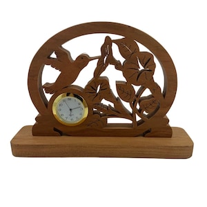 May include: A wooden clock with a hummingbird and floral design. The clock is made of light brown wood and has a white face with black numbers. The hummingbird is in flight and is carved in detail. The floral design is also carved in detail and features leaves and flowers.