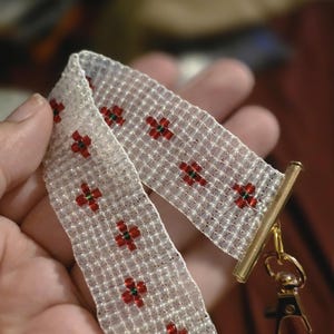 May include: A woven beaded bracelet featuring a translucent white base with a repeating pattern of small red and green flower-like motifs. The bracelet is finished with gold-toned metal end bars and a matching gold-toned lobster claw clasp.