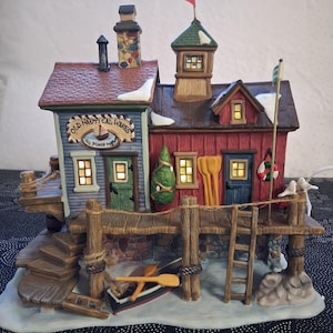 May include: A nautical-themed ceramic village scene with two buildings, a pier, and a small boat. The buildings are painted blue and red, with the text "OLD NAUTICAL WARES" on the blue building. A small Christmas tree is visible. The scene includes a ladder, a flag, and decorative elements.