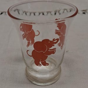 May include: Clear glass shot glass with a flared top and a small base. The glass is decorated with three orange elephant silhouettes. The elephants are in different poses, adding a playful touch to the design.