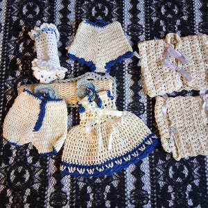 May include: Handmade crochet baby clothing set in cream and blue. The set includes a dress, shorts, a bonnet, a cape, and two pairs of booties. The items are arranged on a black and white lace fabric.