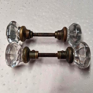 May include: Two antique glass door knobs with brass hardware. The clear, faceted knobs are connected by a dark metal rod. The brass components show signs of age, adding to the vintage aesthetic. These knobs are a classic choice for interior doors.