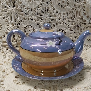 Puede incluir: Una tetera azul y dorada con un diseño floral y un platillo a juego. La tetera tiene una tapa y un pico. El platillo tiene un diseño floral.