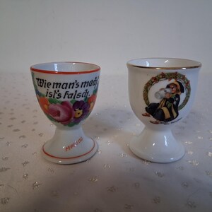 May include: Two white ceramic egg cups. One cup has a floral design with the text "Wie man's mad ist's falsch." The other cup features a figure drinking from a mug, surrounded by a wreath.