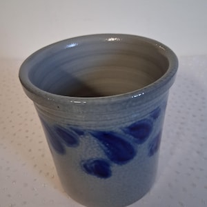 May include: A cylindrical stoneware container with a light gray base and a dark blue floral pattern. The rim is slightly angled, and the interior is a darker shade of gray. The container is likely handmade.