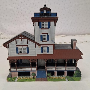 May include: A miniature house model with a white exterior, brown trim, and blue shutters. The model features a porch, a tower, and a brick chimney. The house is decorated with small details like windows and a front door. The model is set on a green base with yellow flowers.