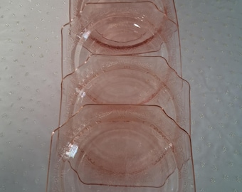 May include: A stack of four pink glass plates with a scalloped edge and a floral pattern. The plates are arranged in a vertical stack, with the top plate slightly offset. The plates are made of a translucent pink glass.