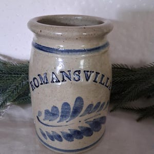 May include: A cylindrical stoneware crock with a speckled beige finish, featuring a blue hand-painted leaf motif and the word ROMANSVILLE embossed in blue lettering. The vessel has a thick, rounded rim and a blue band encircling the top and base.