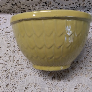 Puede incluir: Un cuenco mezclador de cerámica amarilla con un diseño texturizado y festoneado. El cuenco tiene un borde ancho y un acabado brillante. El fondo es un tapete de ganchillo color crema.