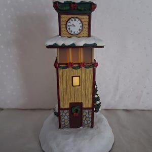 May include: A decorative clock tower figurine featuring a yellow wood-textured facade, a stone-patterned base, and a white snow-covered roof. The tower includes a clock face with Roman numerals, a small illuminated window, and a dark door with a wreath.