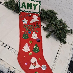 May include: A handmade red Christmas stocking with the name "AMY" in green letters. The stocking is decorated with felt Christmas trees, a reindeer, stars, and the words "Joyeux Noel". Small bells and sequins add festive accents.