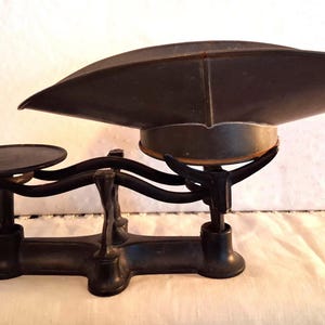 May include: A black cast iron balance scale featuring a wide, shallow, curved metal weighing pan on one side and a smaller, flat, circular platform on the other. The scale has a central support structure with curved arms connecting to the platforms.