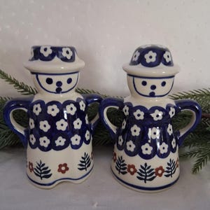 May include: Two ceramic shakers shaped like figures wearing hats, decorated with a blue and white floral pattern and small red flower accents. Each figure features two side handles and a smooth, glossy finish with a painted face design.