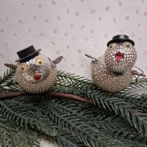 May include: Two round, silver-colored bird ornaments with textured, dimpled surfaces. Each bird features a small black top hat, prominent round eyes, and a red beak. The birds are perched on a green, artificial pine branch with needle-like foliage.
