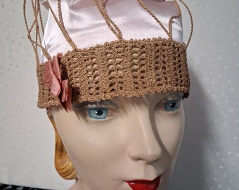 May include: A vintage-style hat with a light pink satin crown and a brown crocheted brim. The hat features a delicate net-like design and a small pink fabric flower. This elegant headwear is perfect for special occasions.