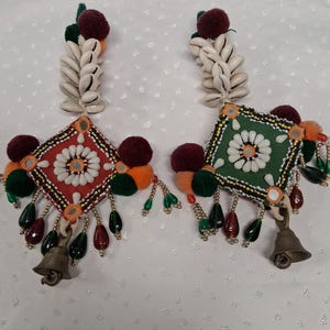 May include: A pair of dangling earrings featuring a diamond-shaped fabric base in red and green. Each earring is adorned with white cowrie shells, small mirrors, colorful pom-poms, green and red teardrop beads, and a small brass bell at the bottom.