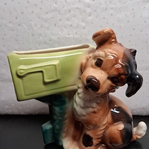 May include: A ceramic dog figurine with a mailbox planter. The dog is brown, black, and white, with a glossy finish. The mailbox is light green. The figurine is a decorative item.