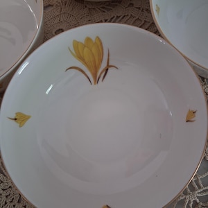 May include: White ceramic bowls with a gold rim and a yellow floral design. The bowls have a shallow, wide shape and are decorated with a large yellow flower and smaller floral accents. The bowls are likely part of a dinnerware set.