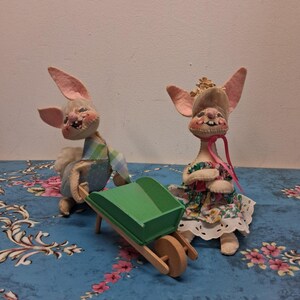 May include: Two whimsical bunny dolls, one pushing a green wheelbarrow. The other bunny wears a floral dress and a crown. The dolls have pink ears and are set on a blue floral patterned surface.