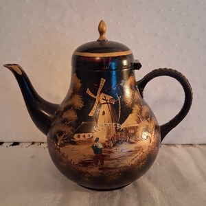 May include: A black teapot featuring a hand-painted gold landscape scene of a windmill, a small house, and a person walking. The teapot has a curved spout, a textured handle, and a lid topped with a small gold-colored finial knob.