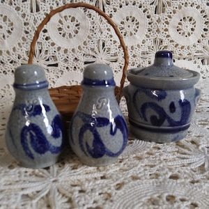 May include: A set of three blue and gray ceramic salt and pepper shakers with a blue and white design. The shakers are in a wicker basket on a white crocheted tablecloth.
