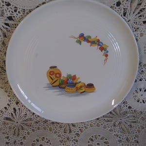 May include: White ceramic plate with a colorful fruit and pottery design. The plate features a curved arrangement of various fruits and vegetables, along with several small, decorative pots. The plate has a simple, elegant design.