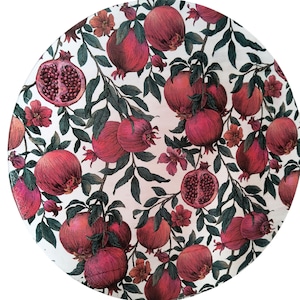 Puede incluir: Plato decorativo redondo con diseño de granadas. El plato presenta un fondo blanco con un patrón de granadas rojas, hojas verdes y pequeñas flores naranjas. El texto "De_olié" está escrito en cursiva en la parte inferior.