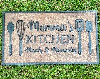 May include: Wooden kitchen sign with the words "Momma's Kitchen Meals & Memories" in a decorative font. The sign features embossed kitchen utensils: a spoon, whisk, spatula, and turner. The sign has a dark brown frame and a light brown interior.