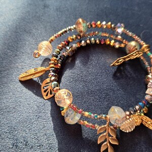 Könnte beinhalten: Ein mehrsträngiges Armband mit goldfarbenen Blatt- und Federanhängern. Das Armband hat eine Mischung aus bunten Perlen, darunter schillernde und klare Perlen. Das Armband liegt auf einer dunkelblauen Oberfläche.