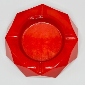 K&ouml;nnte beinhalten: Ein leuchtend roter, facettierter, geometrischer Aschenbecher mit einem kreisf&ouml;rmigen Becken in der Mitte. Die Oberfl&auml;che hat ein glattes, gl&auml;nzendes Finish mit feinen, schimmernden Partikeln, die f&uuml;r ein lebendiges und reflektierendes Aussehen sorgen.