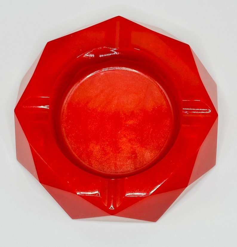 K&ouml;nnte beinhalten: Ein leuchtend roter, facettierter, geometrischer Aschenbecher mit einem kreisf&ouml;rmigen Becken in der Mitte. Die Oberfl&auml;che hat ein glattes, gl&auml;nzendes Finish mit feinen, schimmernden Partikeln, die f&uuml;r ein lebendiges und reflektierendes Aussehen sorgen.
