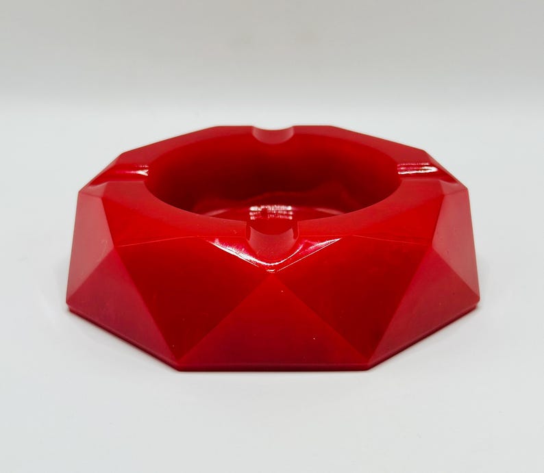 K&ouml;nnte beinhalten: Ein leuchtend roter Aschenbecher mit facettiertem, geometrischem Au&szlig;endesign. Das Objekt verf&uuml;gt &uuml;ber ein kreisf&ouml;rmiges zentrales Becken, das von mehreren abgewinkelten, flachen Oberfl&auml;chen umgeben ist, die eine scharfe, mehrseitige Form bilden.