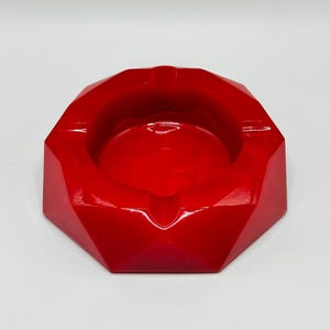 K&ouml;nnte beinhalten: Ein leuchtend roter Aschenbecher mit facettiertem, geometrischem Design, das mehrere kantige Seiten und eine glatte, gl&auml;nzende Oberfl&auml;che aufweist. Das kreisf&ouml;rmige Becken ist von einem dicken, mehrseitigen Rand mit integrierten Ablagekerben umgeben.