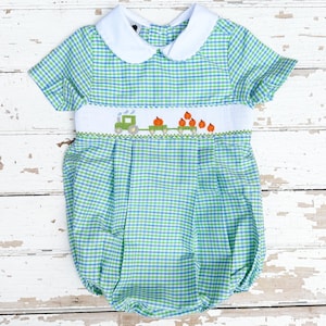May include: A baby romper in a blue and green gingham pattern. The romper features a white collar and a smocked panel with a train carrying pumpkins. The short-sleeved romper is designed for infants and toddlers.