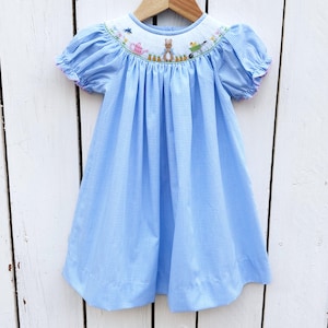 May include: A light blue gingham dress with short, puffed sleeves and a white embroidered yoke. The yoke features a garden scene with a bunny, watering cans, and a wheelbarrow. The sleeves are trimmed with pink. The dress is hanging on a wooden hanger.