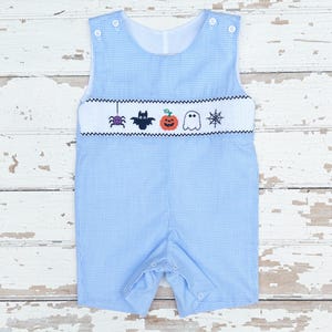 May include: Light blue and white checkered romper with a Halloween-themed embroidered design. The design includes a spider, bat, jack-o'-lantern, ghost, and spiderweb. The romper has button closures and a white collar.
