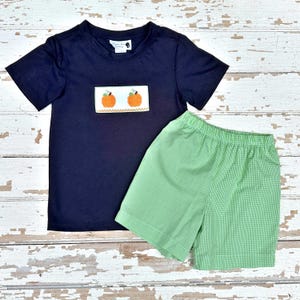 May include: Navy blue short-sleeved shirt and green gingham shorts set. The shirt features an embroidered patch with two orange pumpkins. The shorts are a light green color with a small checkered pattern. The set is likely for children.