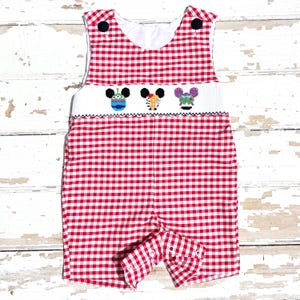 May include: Red and white gingham romper with embroidered character faces. The romper features a white bib with three colorful character faces and navy blue buttons on the straps. The shorts have button closures.