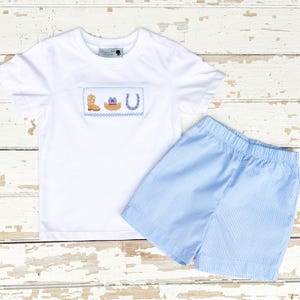 May include: A white short-sleeved t-shirt and blue gingham shorts set. The shirt features a stitched design of a cowboy boot, hat, and horseshoe. The shorts have an elastic waistband.