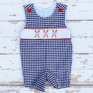 May include: A navy and white gingham romper with red button accents. The romper features a white panel with embroidered baseballs and red trim. The sleeveless design is perfect for warm weather and is a classic style.