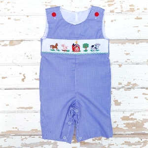 May include: Blue and white gingham romper with red button closures. The romper features an embroidered farm scene with a horse, pig, barn, tree, and cow. The romper has a white collar and is made of a lightweight fabric.