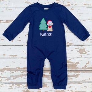 May include: Navy blue long-sleeved baby romper with a Christmas tree and dog applique. The dog wears a Santa hat. The name "WALKER" is embroidered below the applique. The romper has snap closures at the bottom.