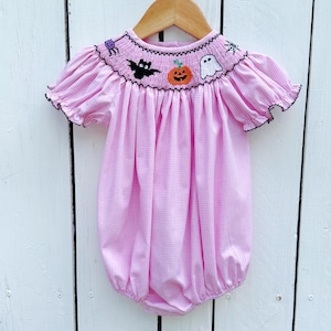 May include: Pink gingham baby romper with short sleeves and a round neckline. The neckline is embroidered with Halloween-themed designs, including a spider, bat, jack-o'-lantern, and ghost. The sleeves and neckline are trimmed with black. The romper is hanging on a wooden hanger.