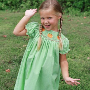 May include: A young girl wearing a green gingham dress with orange pumpkin embroidery on the neckline. The dress has short, puffed sleeves and a brown trim. The girl has blonde braids and is smiling.