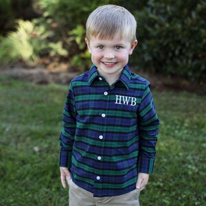 May include: A long-sleeved plaid shirt in shades of green, navy blue, and black. The shirt features white buttons and the embroidered initials "HWB" on the chest. The shirt is worn by a young child.