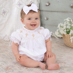 May include: A smiling baby wearing a white romper with gold cross embroidery and a white bow headband. The romper has puff sleeves with lace trim. The baby is sitting on a patterned rug.