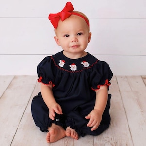 May include: A baby wearing a navy blue romper with red trim and Santa Claus embroidery around the neckline. The baby also wears a red headband with a large bow. The outfit is designed for a baby.